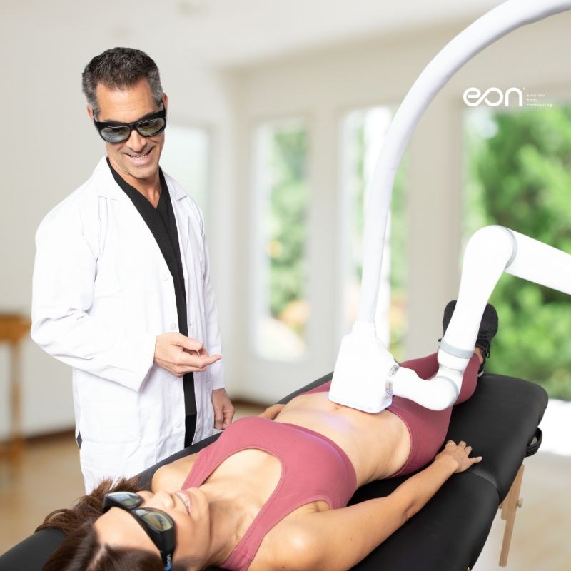 A clinician with glasses speaking with a person reclining beside an EON device.